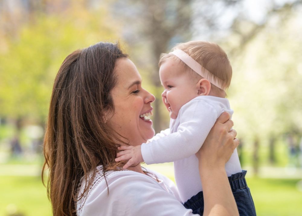 A loving parent lifts a baby up in the air while playing outdoors in a sunny park setting.