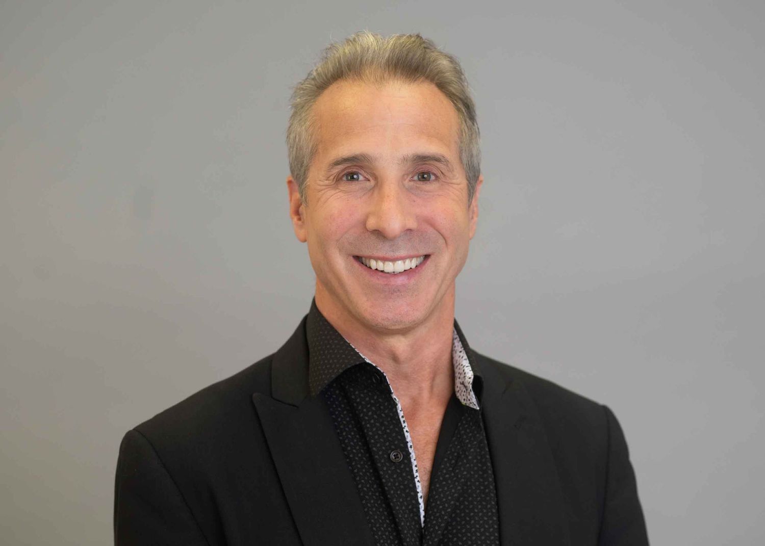 Smiling middle-aged man with gray hair wearing a black blazer and patterned shirt against a gray background.