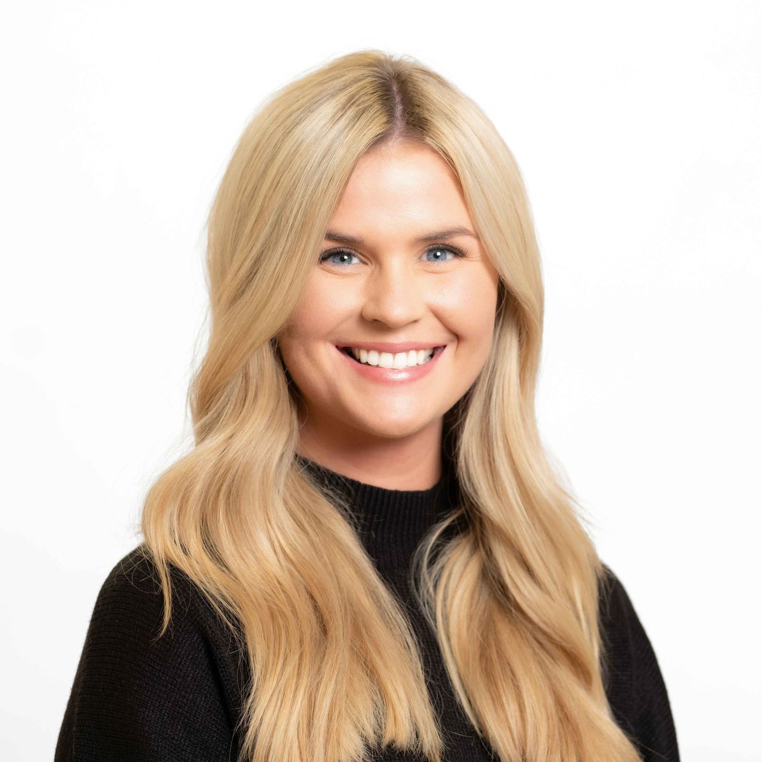 Smiling blonde woman with long wavy hair wearing a black top against a white background.