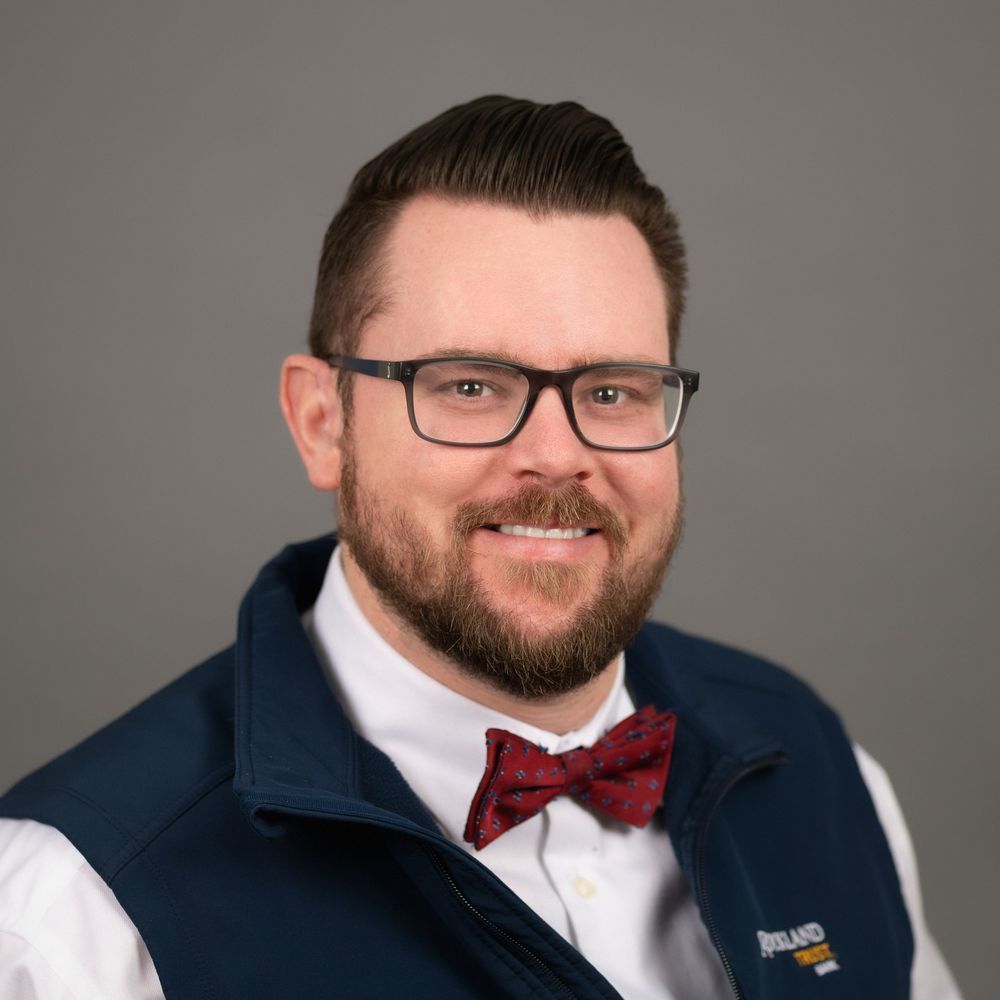 Professional man with beard and glasses wearing a navy vest and red bow tie smiling for a corporate headshot.