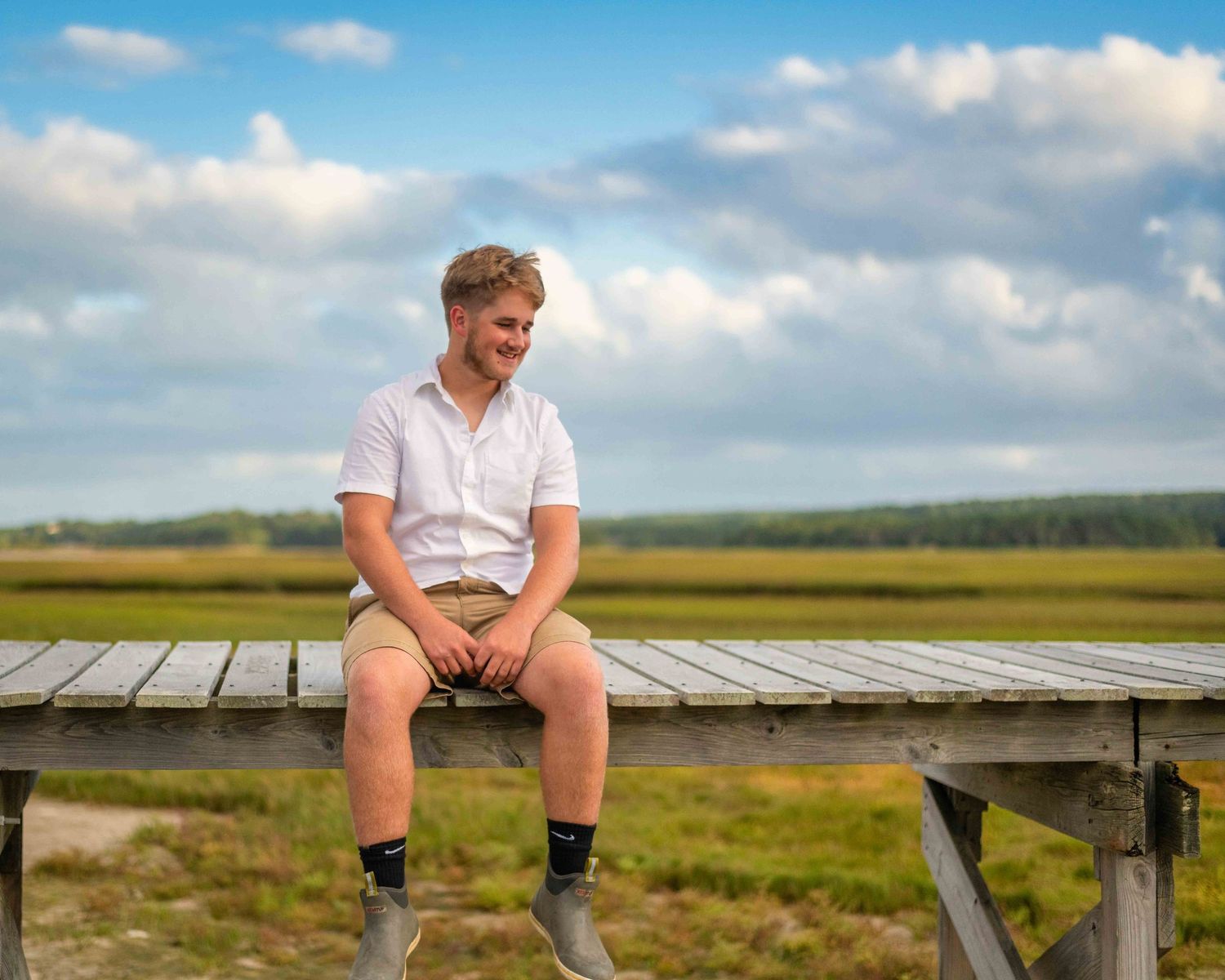 Young man in white shirt and khaki shorts sitting on wooden dock overlooking green marsh under blue sky.