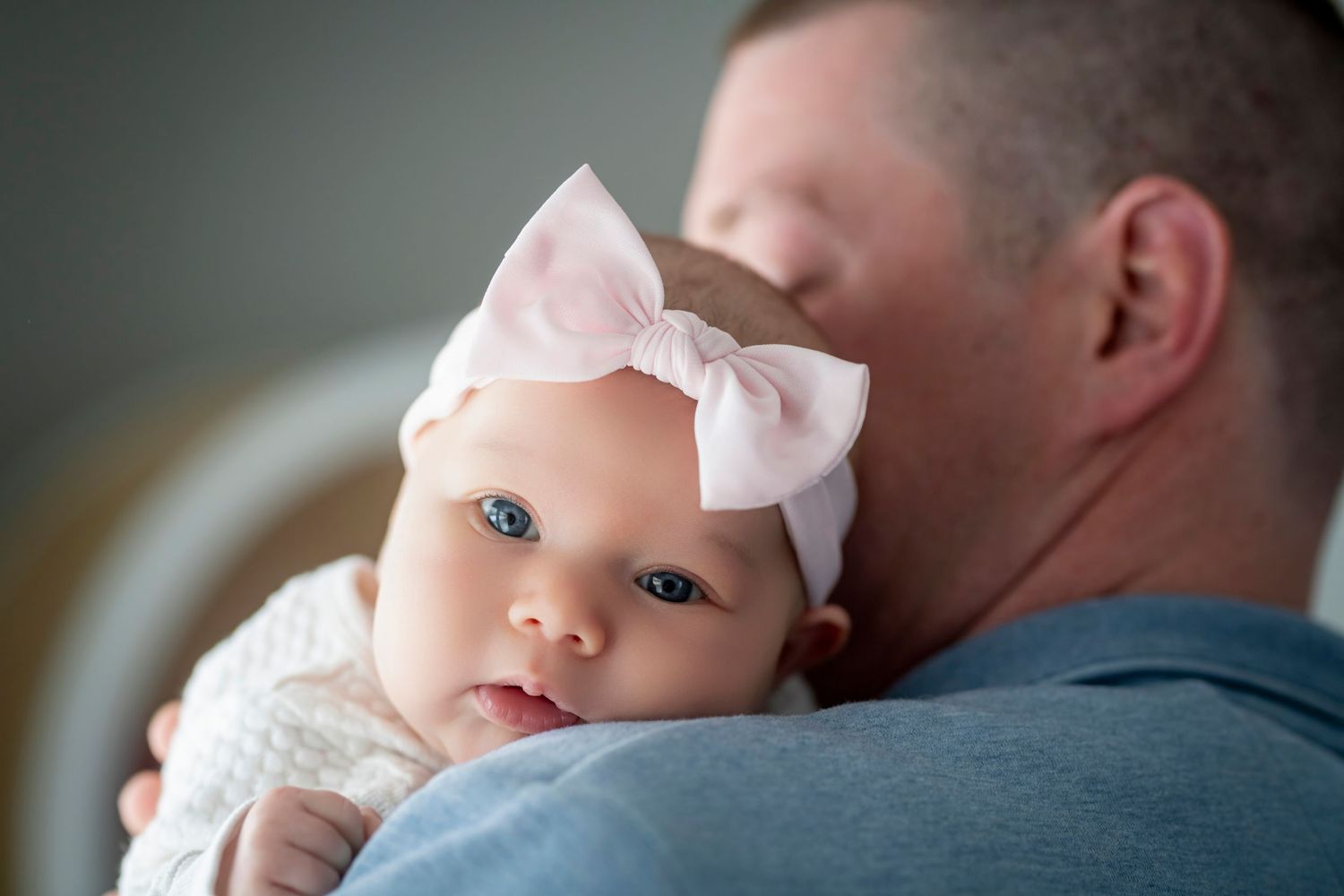 Baby in pink bow wearing white being cradled against someone in blue clothing.