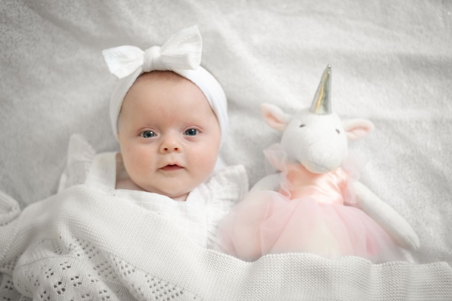Baby in white bow headband lying on blanket next to pink unicorn plush toy.