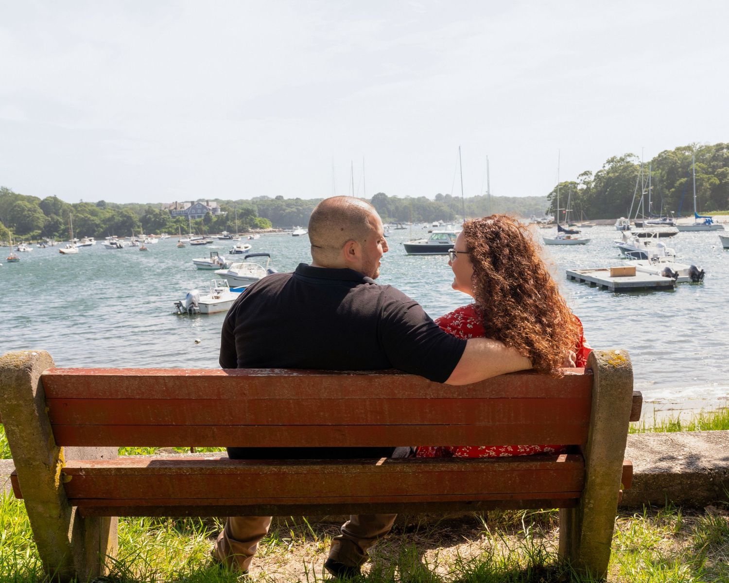 Two people sitting close together on a wooden bench overlooking a harbor with boats.