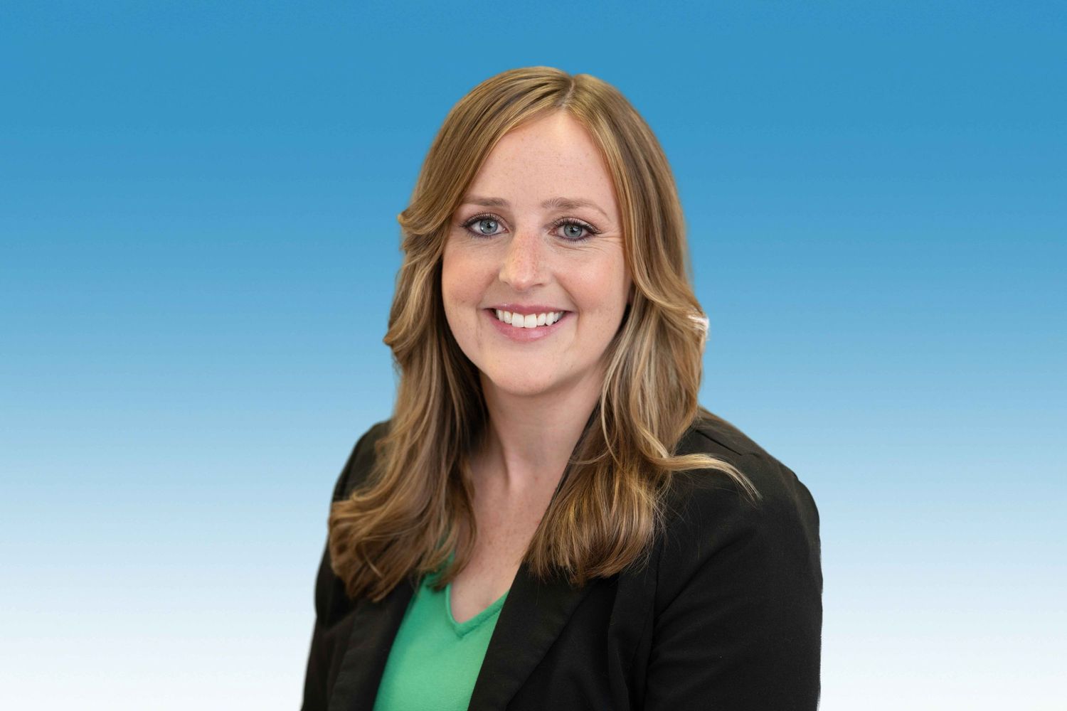 Professional headshot of a smiling woman with long brown hair wearing a black blazer and green top on blue background.