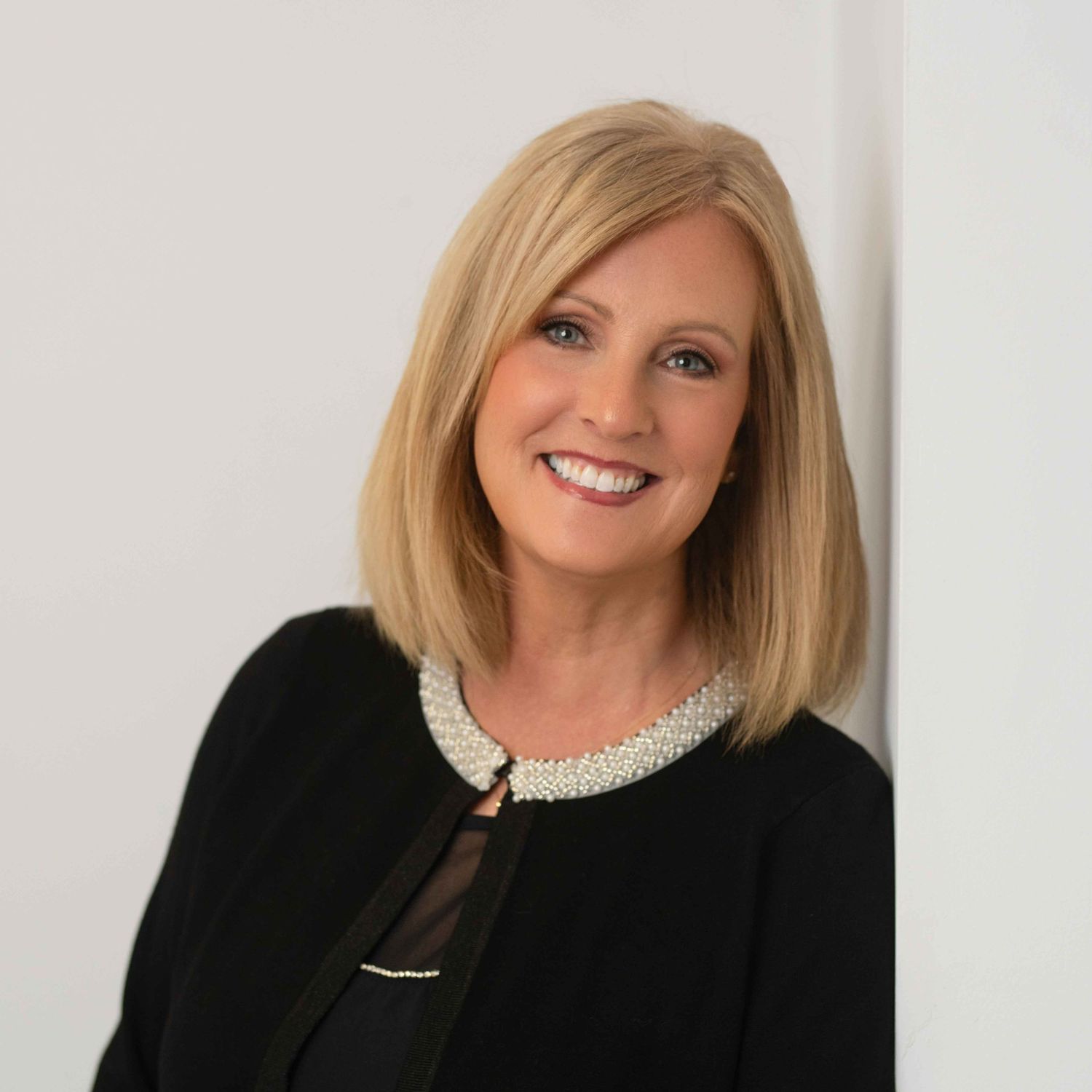 Professional headshot of a smiling blonde woman in a black blazer with pearl neckline detail against a white background.