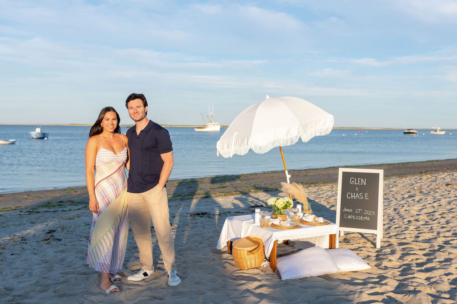 A beach picnic setup with white umbrella, blanket and basket on the sand near the ocean.