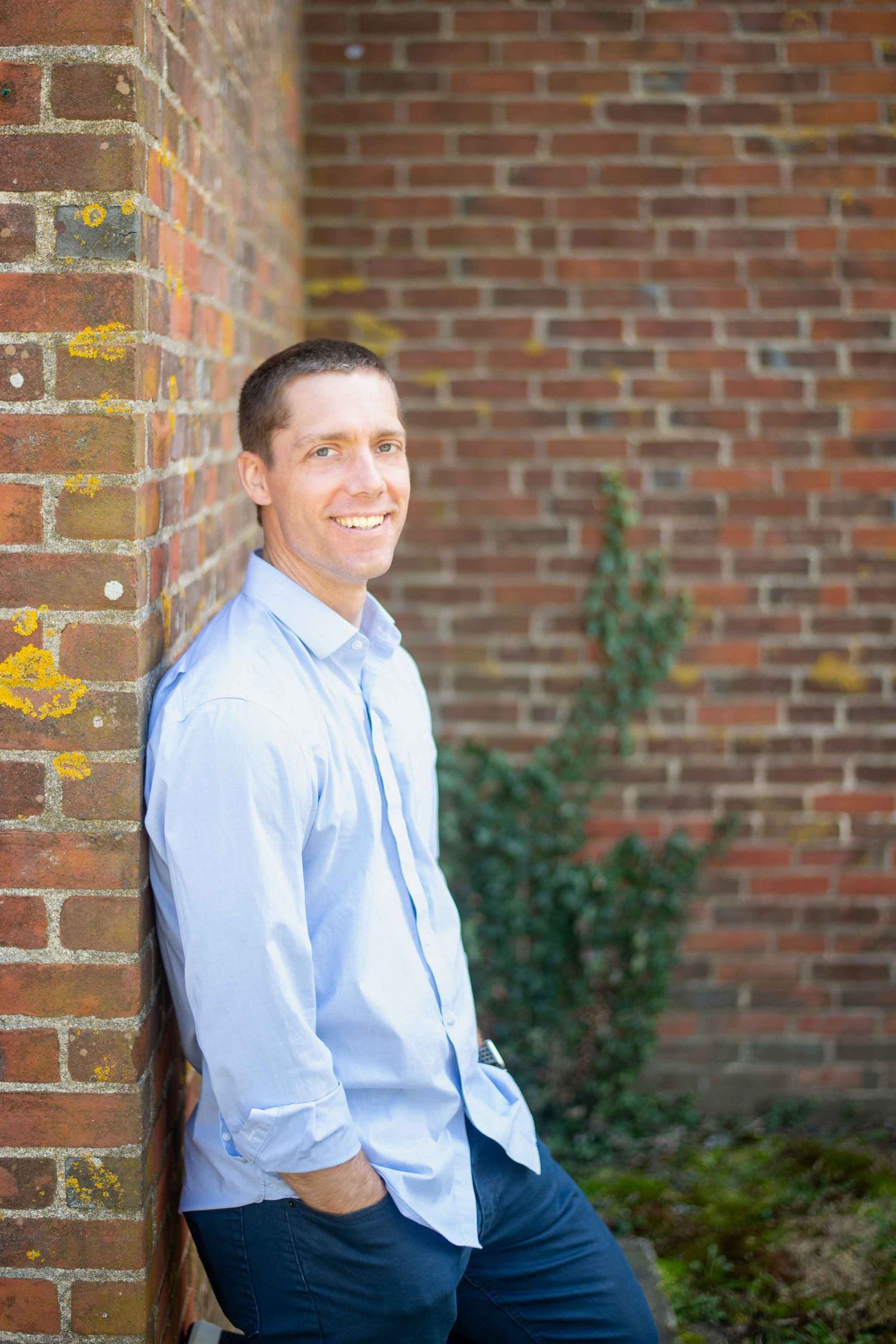 Smiling man in light blue shirt leaning against a brick wall outdoors.