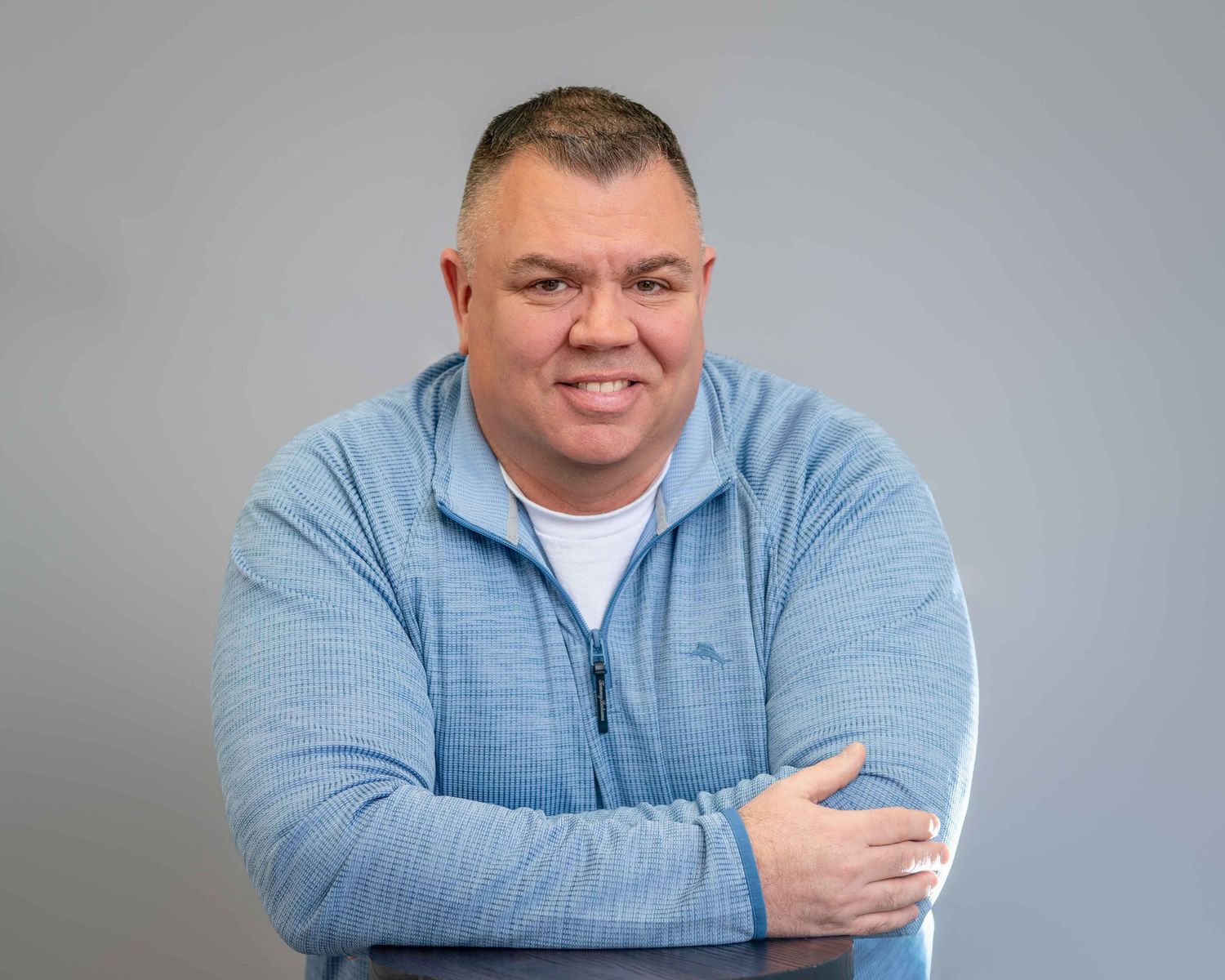Smiling man in blue zip-up hoodie with arms crossed against gray background.