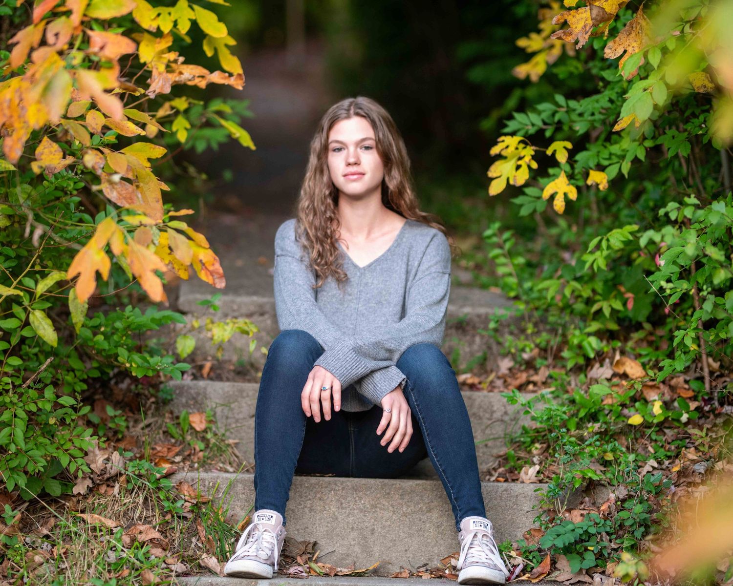 Teen girl with curly hair in gray sweater and jeans sitting on stone steps surrounded by autumn foliage.