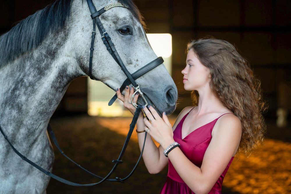 Young woman in red dress tenderly bonding with a gray horse inside a stable.
