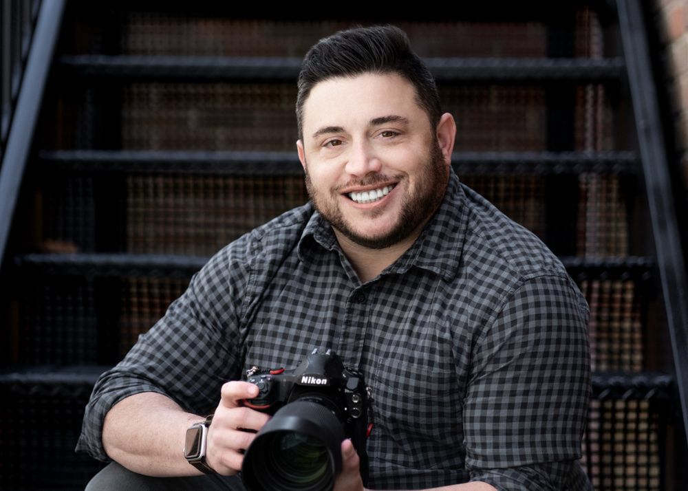 Smiling man holding a Nikon camera while sitting on stairs, wearing a plaid shirt.