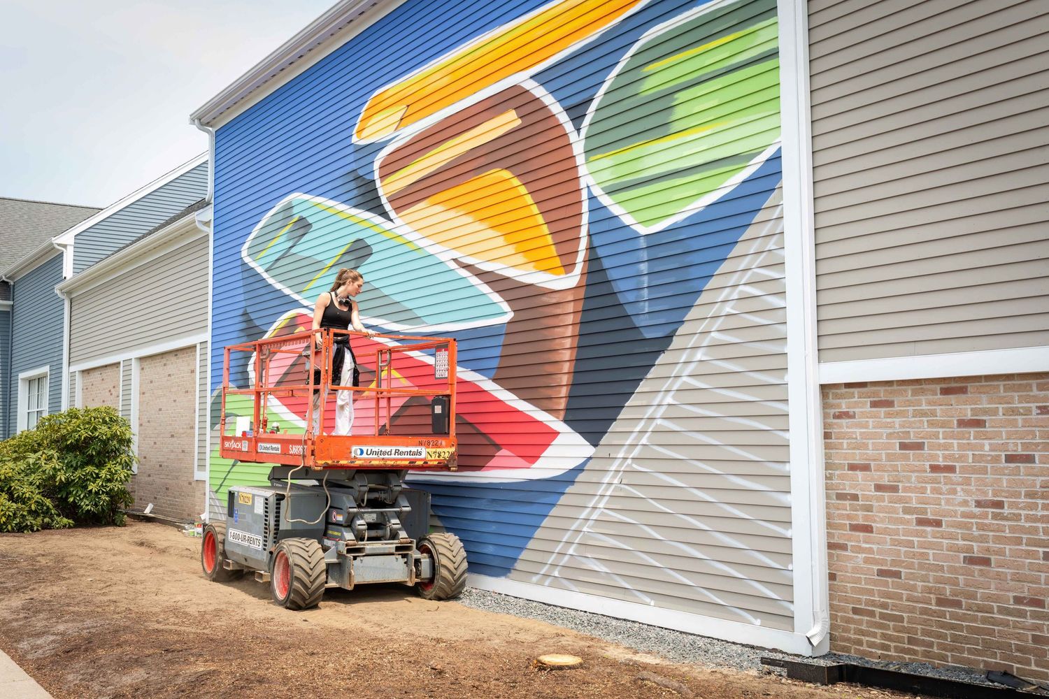Artist on scissor lift painting vibrant colorful mural on exterior brick building wall outdoors.