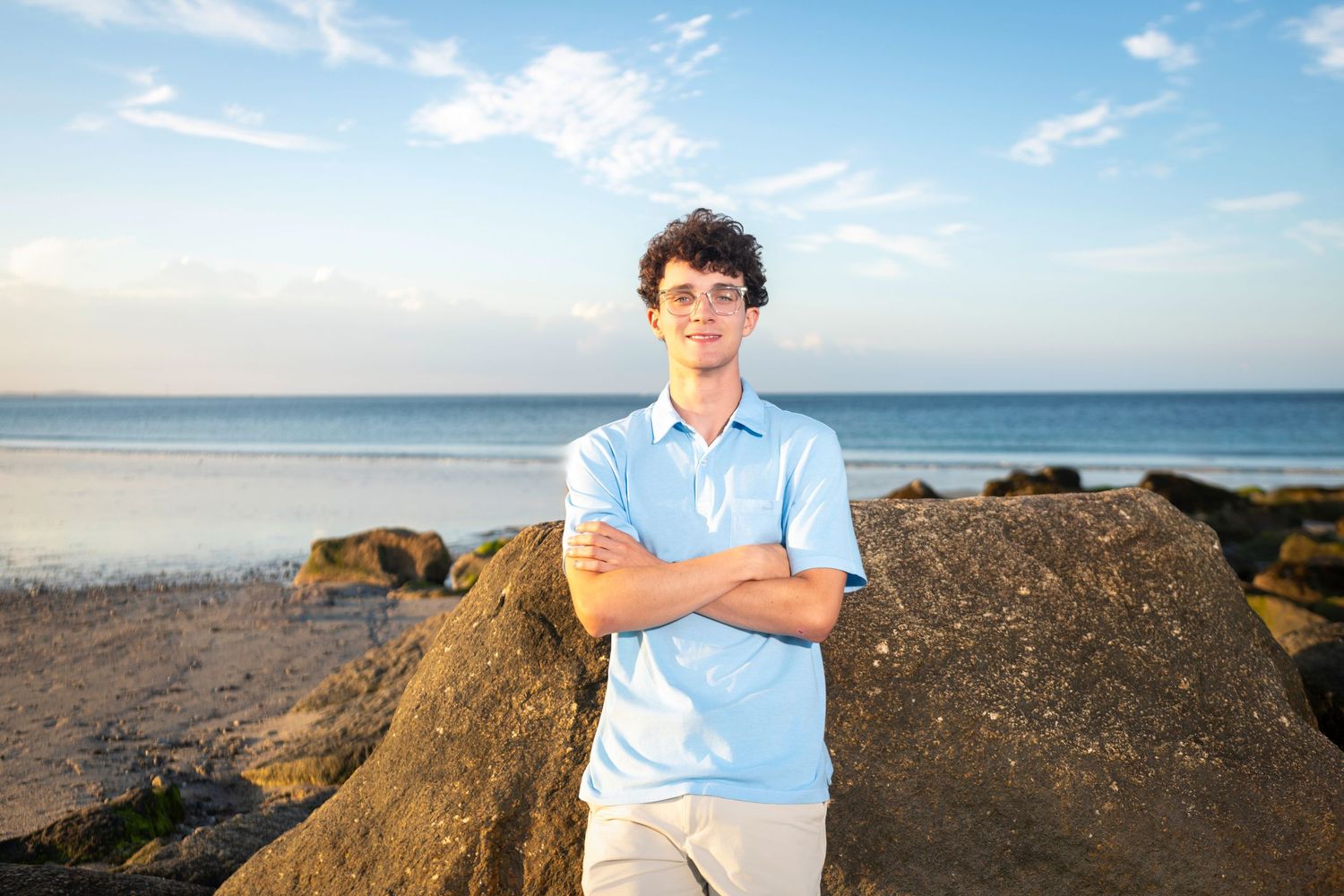 Young man in light blue polo stands arms crossed on rocky beach with ocean horizon at sunset.