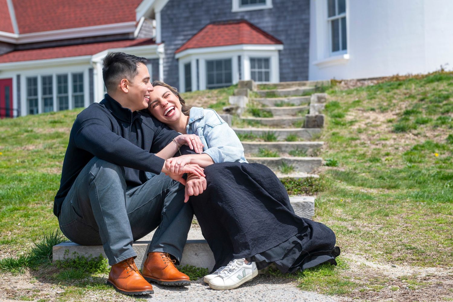 A couple sits together on stone steps near a classic New England coastal home.