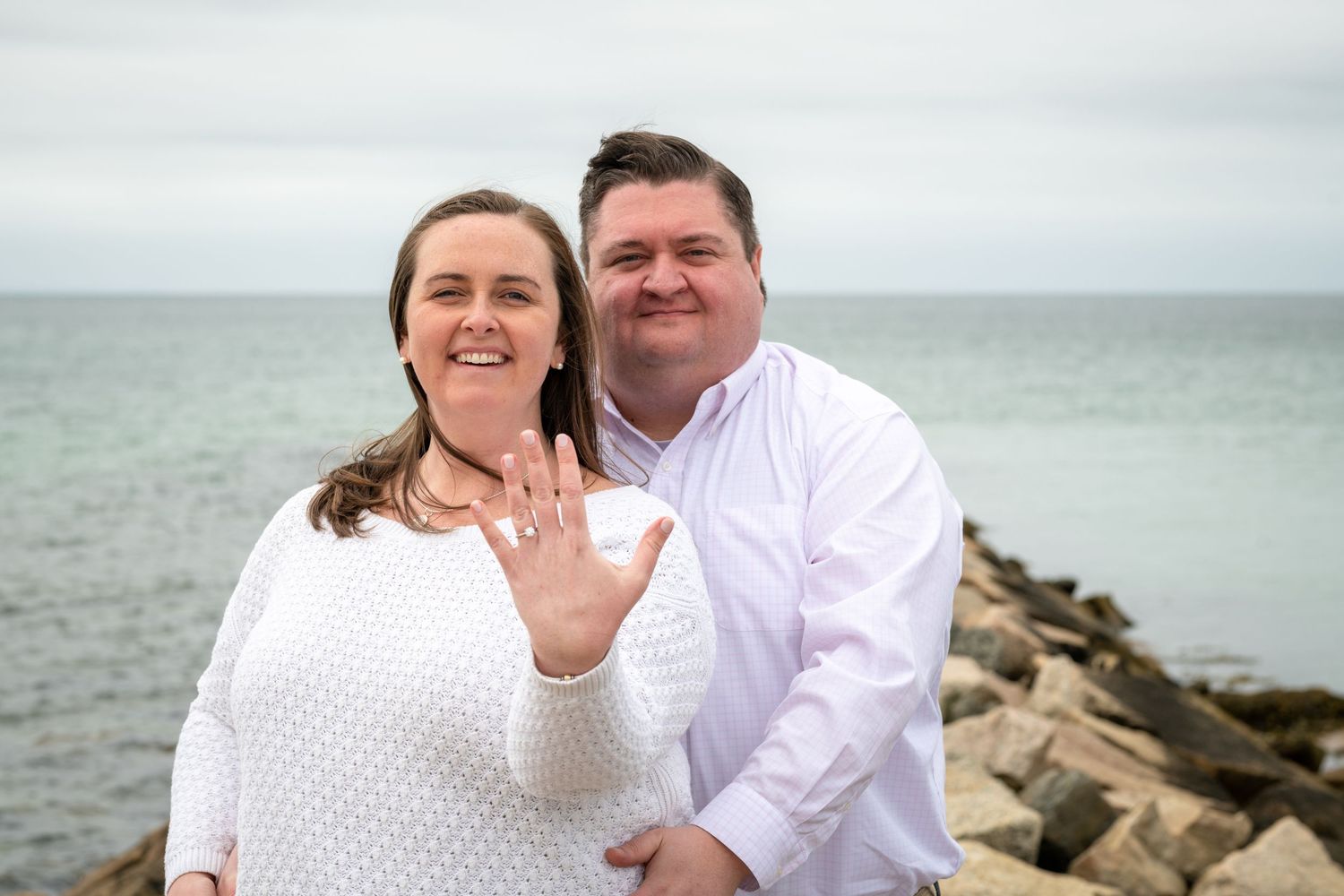 Happy couple showing off engagement ring on rocky beach overlook.
