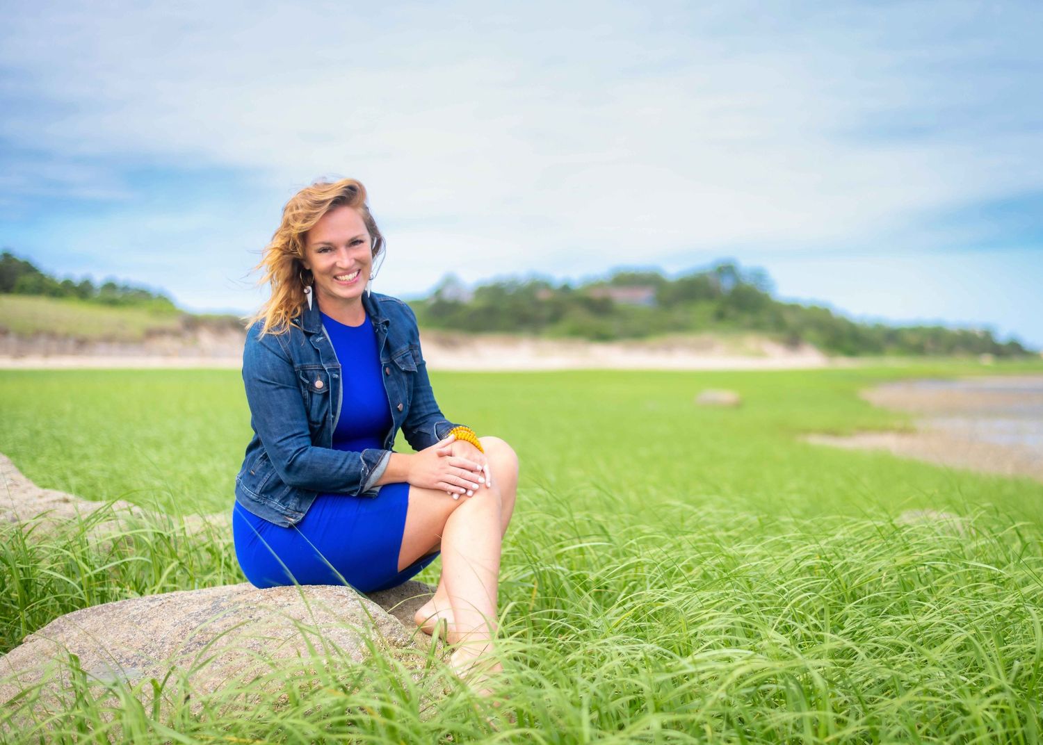 Smiling woman in blue dress and denim jacket sitting on rock in lush green coastal marsh landscape.