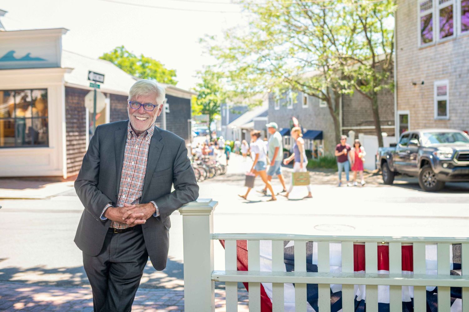 Smiling older man in suit leaning on white fence on sunny small-town street with pedestrians and trees in background.