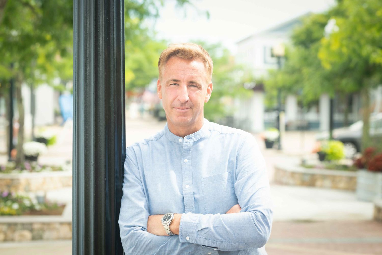 Man in light blue shirt leaning against pole outdoors with arms crossed.
