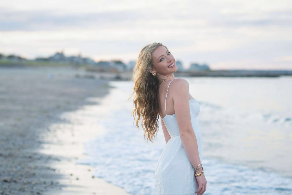 Young woman in white dress smiling on a beach at dusk with ocean waves and coastal town in background.