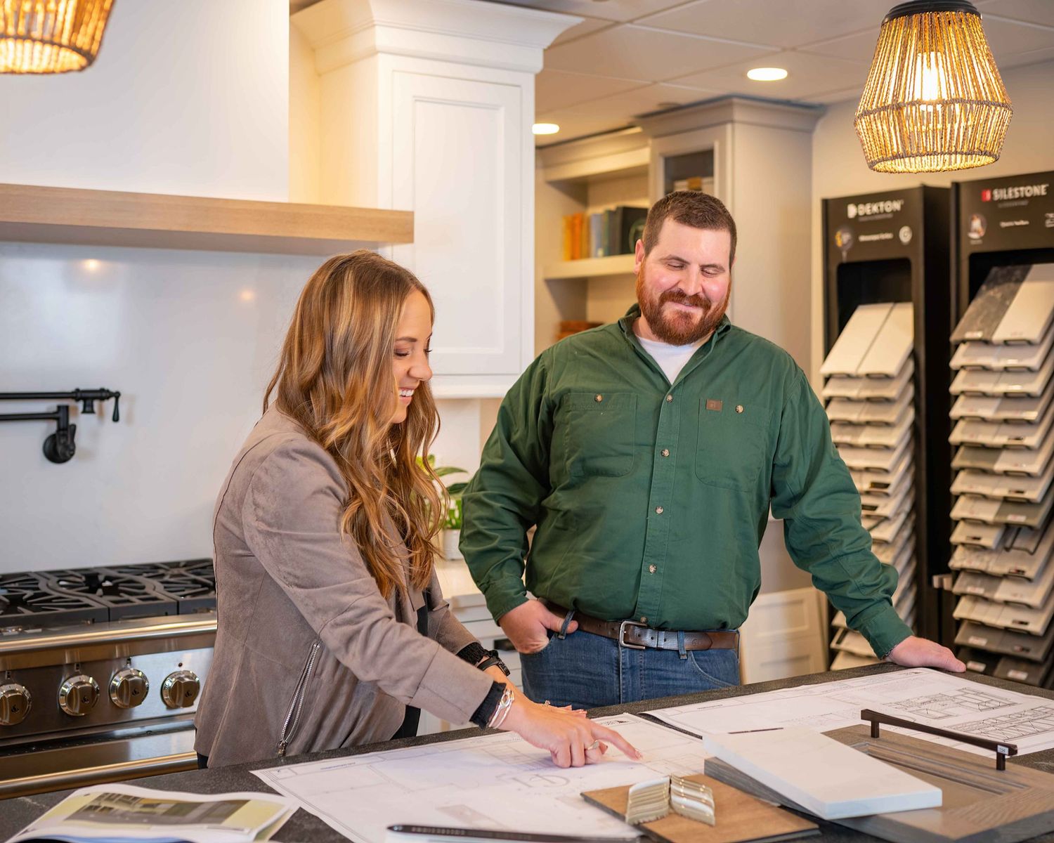 Two people reviewing design plans at a kitchen showroom counter with cabinet and flooring samples displayed.