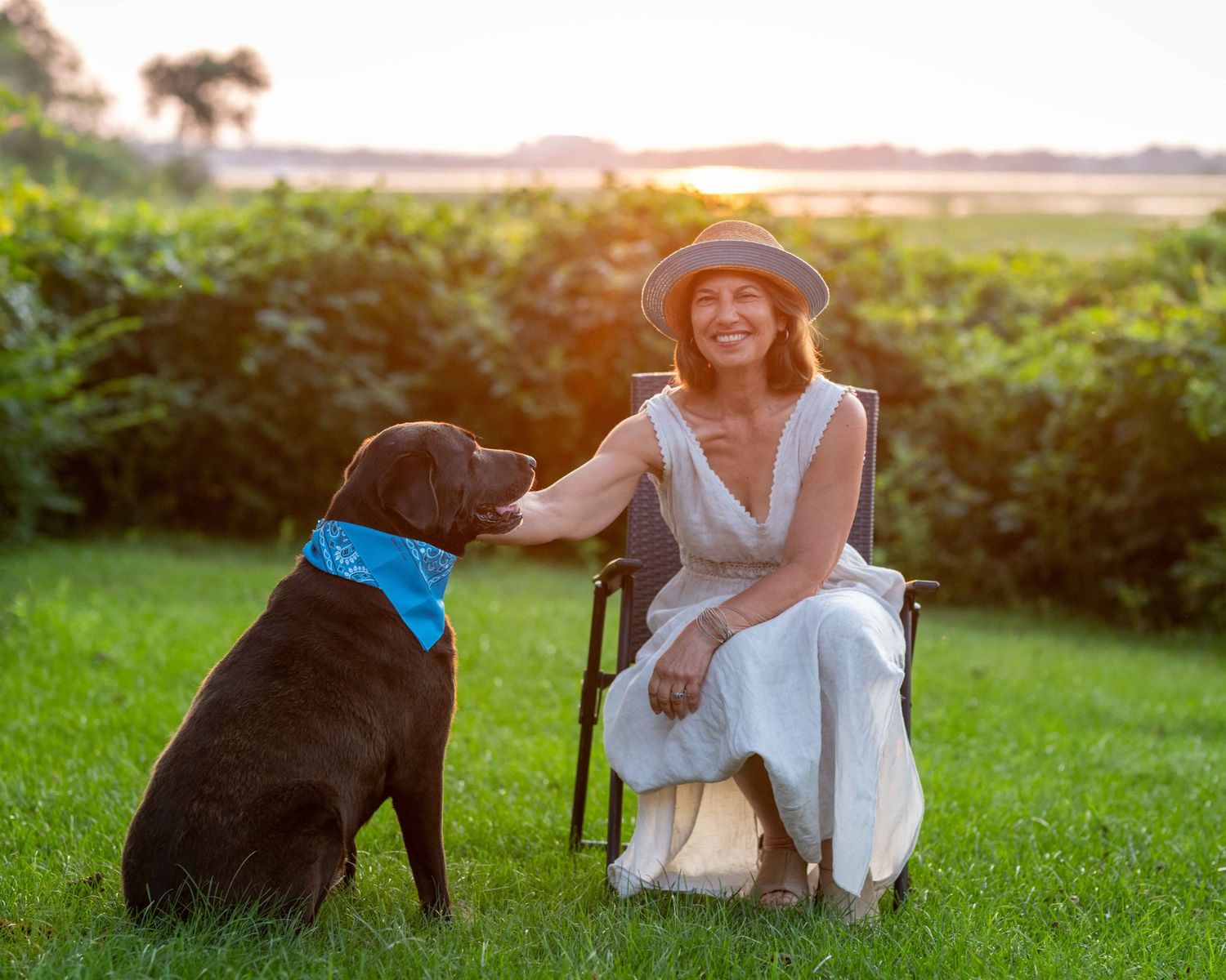 Woman in hat sits in chair petting chocolate lab dog wearing blue bandana on sunny green lawn.