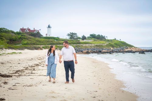 A couple walks hand in hand along a sandy beach with a white lighthouse perched on the coastal cliffs in Maine.