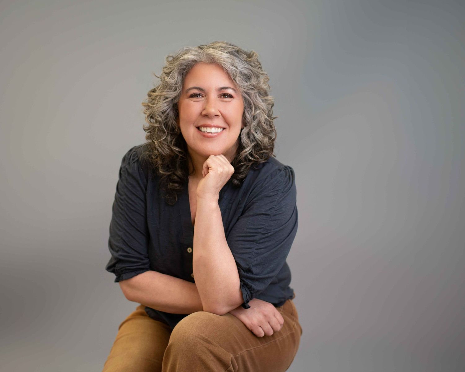 Smiling woman with curly gray hair sitting relaxed with hand on chin against gray backdrop.