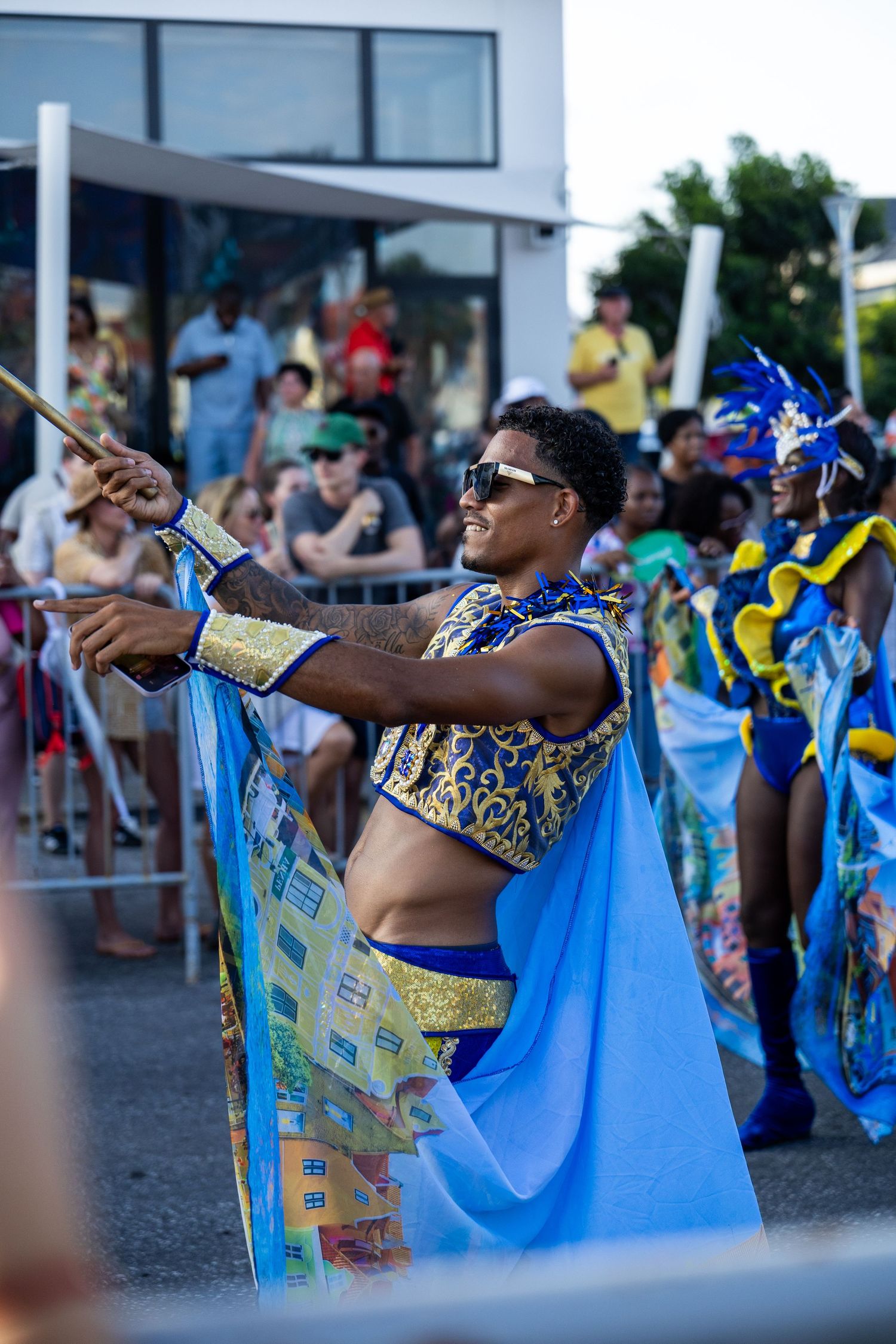 Festival dancer in gold and blue costume performs with batons at street celebration.