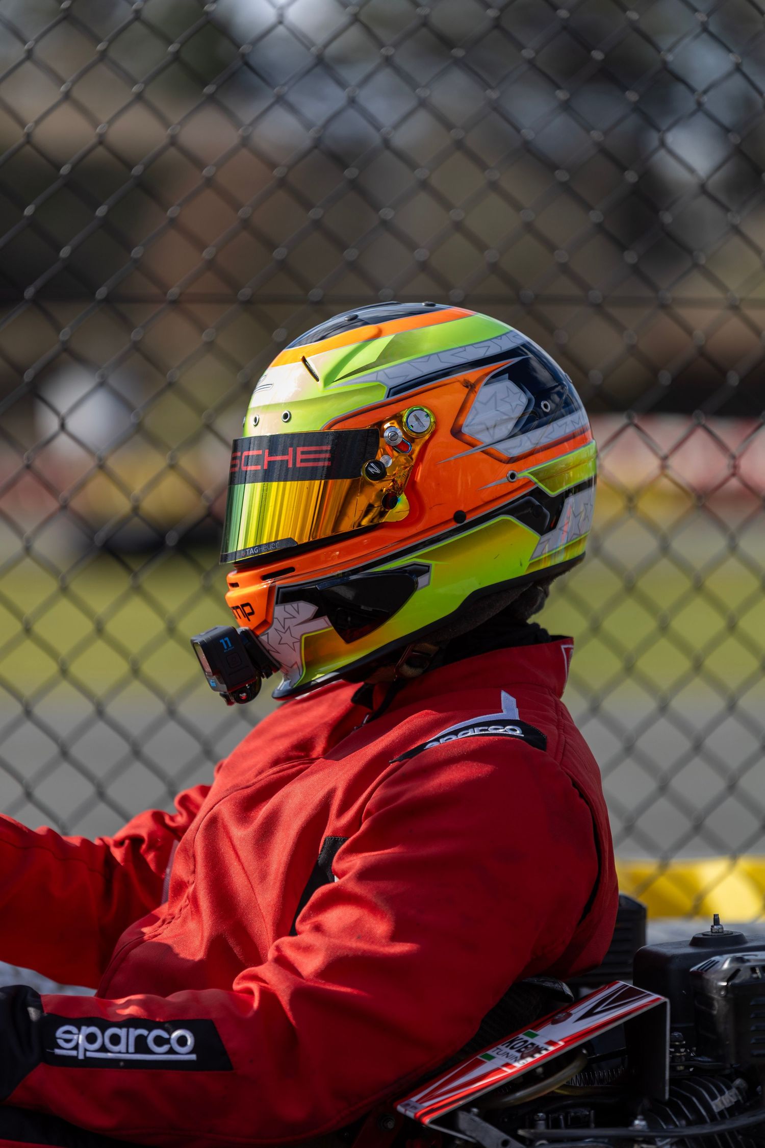 A race car driver wearing a bright neon helmet and red Sparco racing suit sits in their kart against a chain-link fence background.