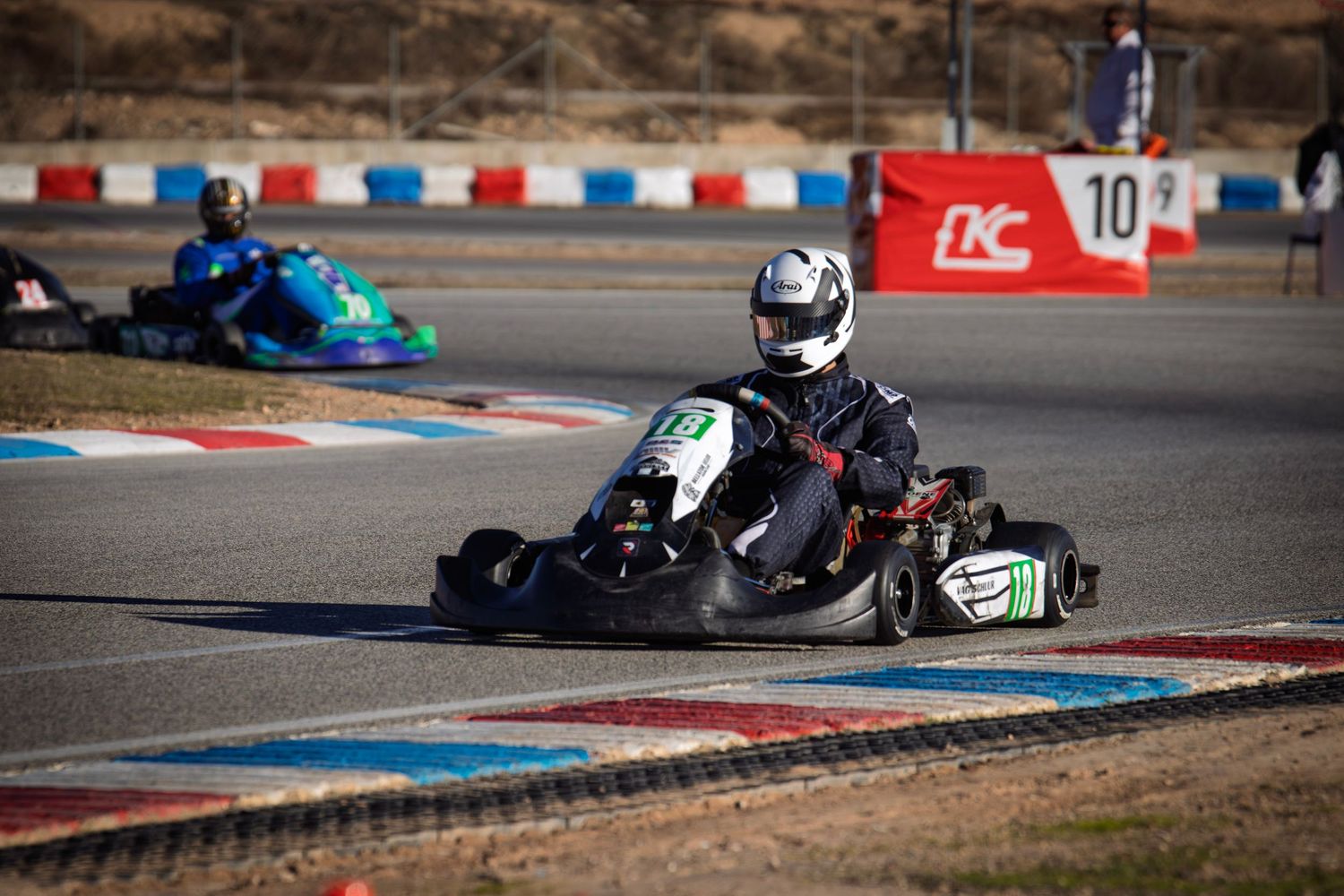 Go-kart in dark livery navigates a corner with blue kart visible in background.