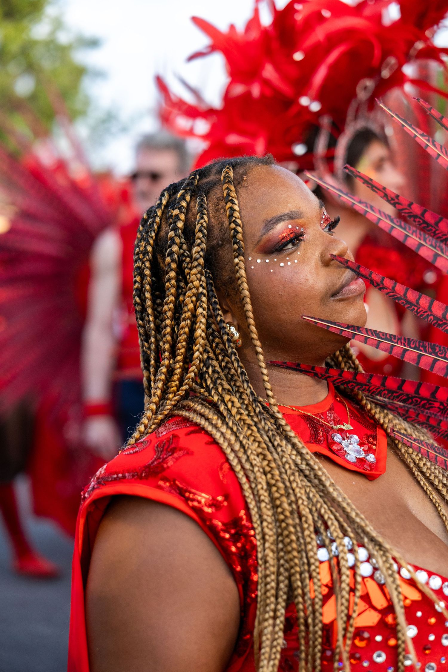 Performer with braided hair and red sequined costume wears an ornate feathered carnival headdress.