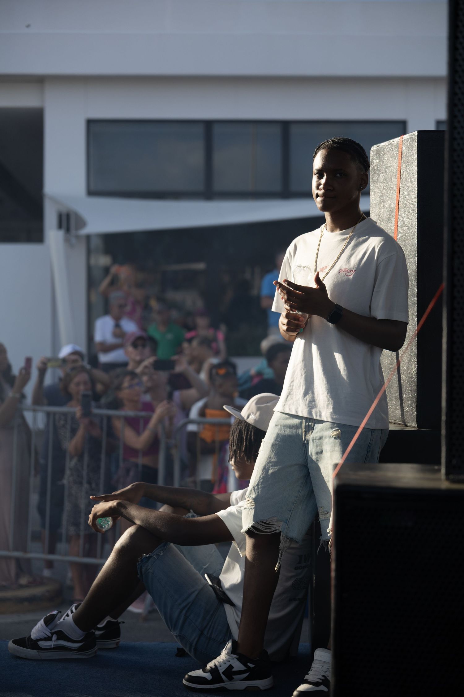 Performer standing on outdoor stage with crowd barriers visible in background.