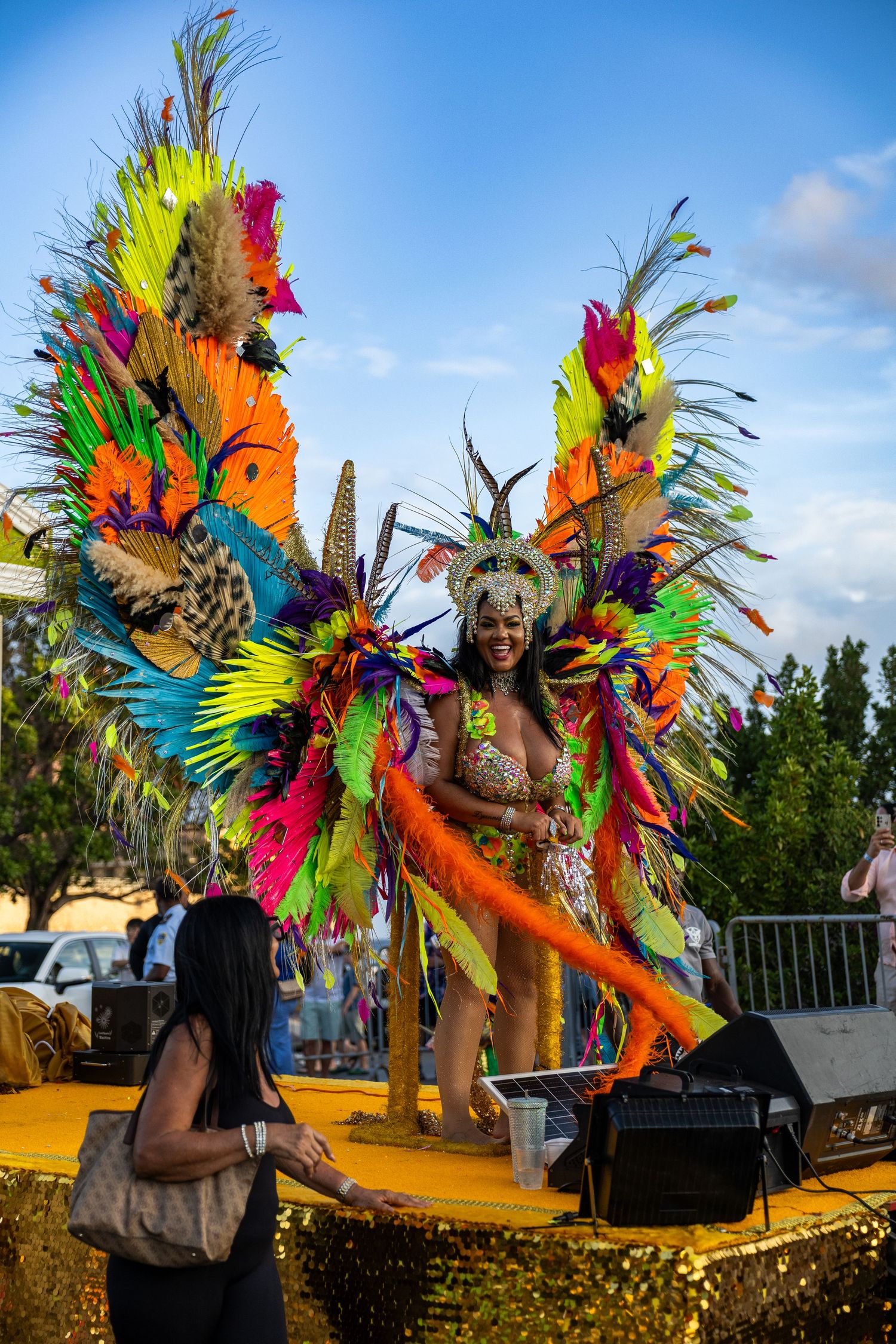 Performers wearing colorful feathered carnival costumes dance against a blue sky backdrop.