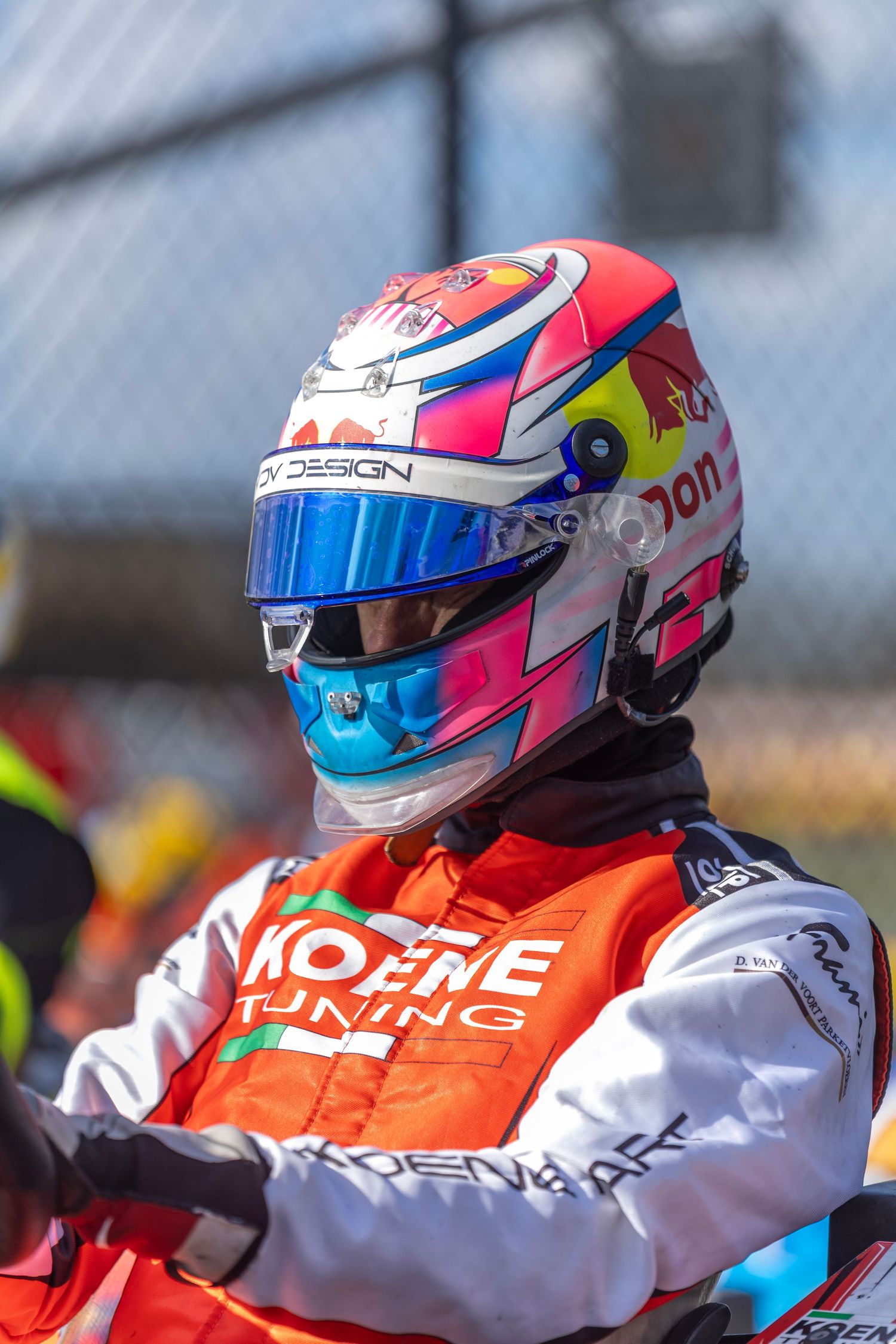 Racing driver wearing orange and white Modene Tuning suit with colorful Red Bull style helmet in bright sunlight.