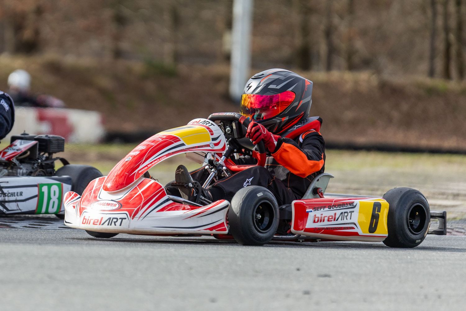 Go-kart racer in red and white kart navigates a turn on outdoor racing track during competition.