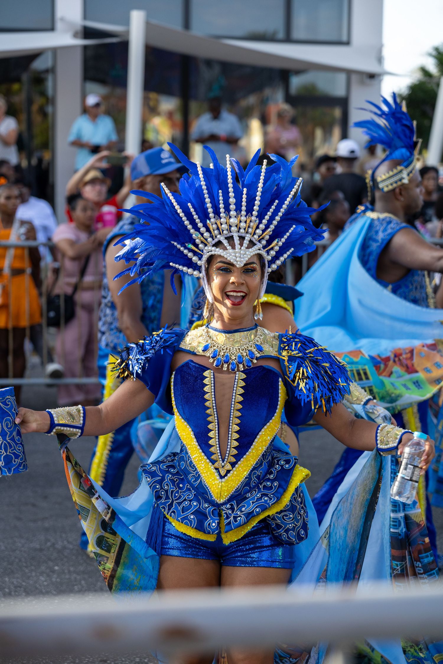 Carnival dancer in vibrant blue feathered costume and headdress performing at a festival celebration.
