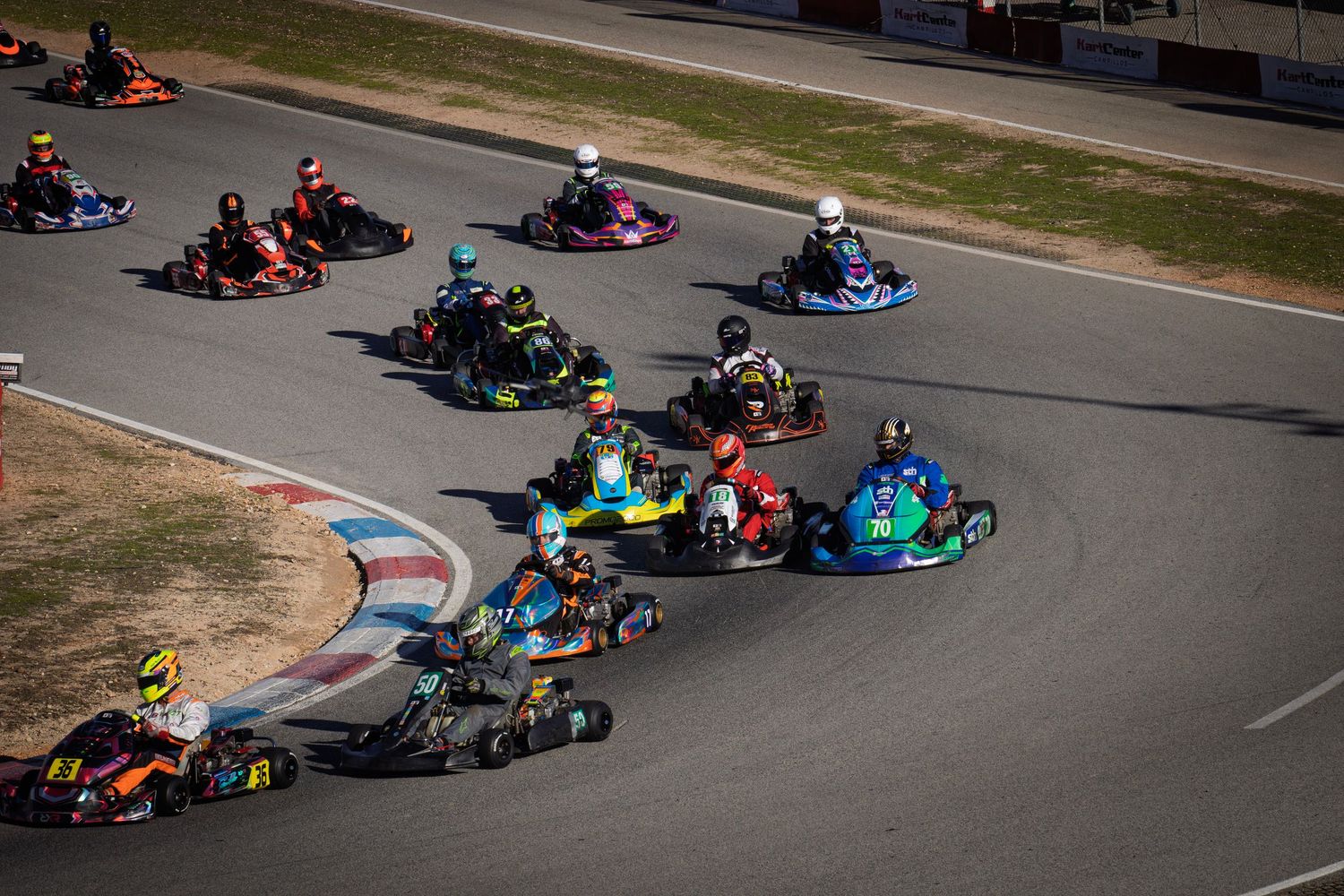 Go-karts racing around a paved track with red and blue curbing during a competitive motorsport event.