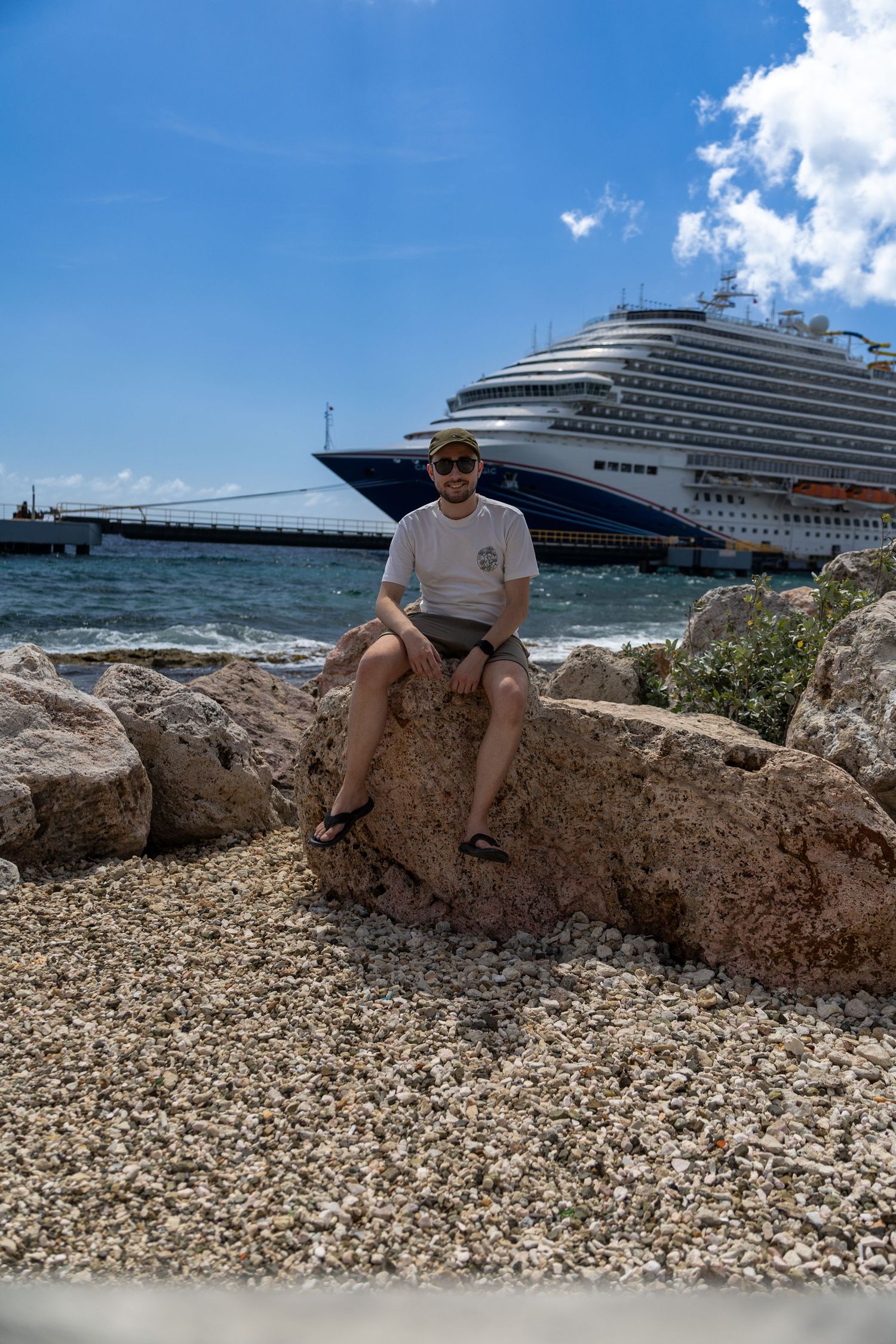 Cruise ship passenger sits on rocky shore with large luxury liner docked in Caribbean port.