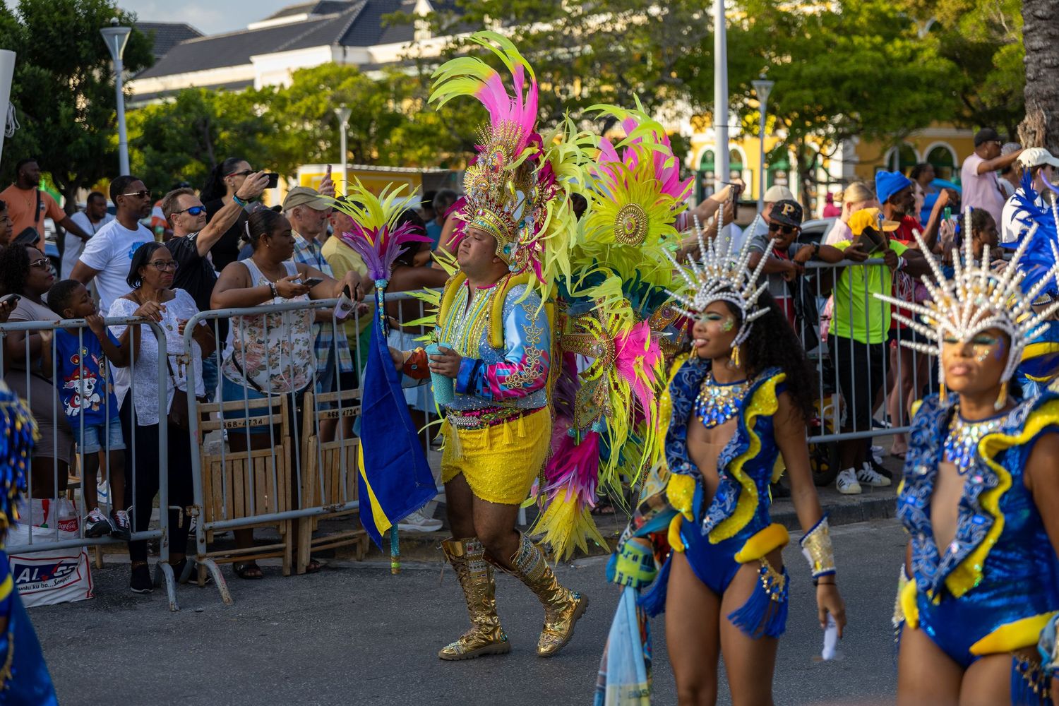 Colorful carnival performers in feathered costumes entertain crowd behind street barriers.