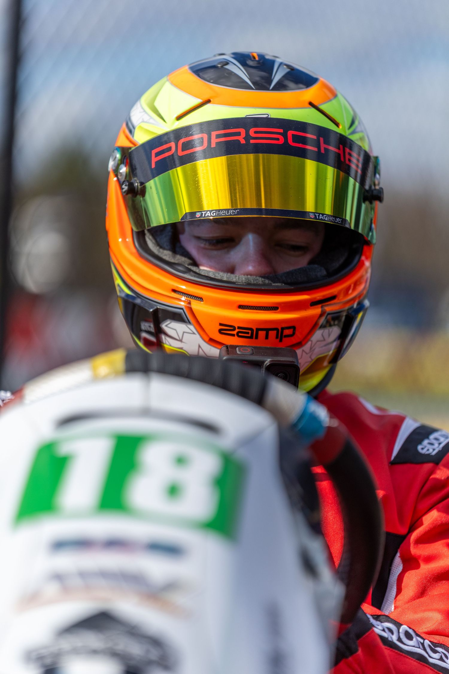 Close-up of a racing helmet with orange and yellow design featuring Porsche branding and reflective visor.