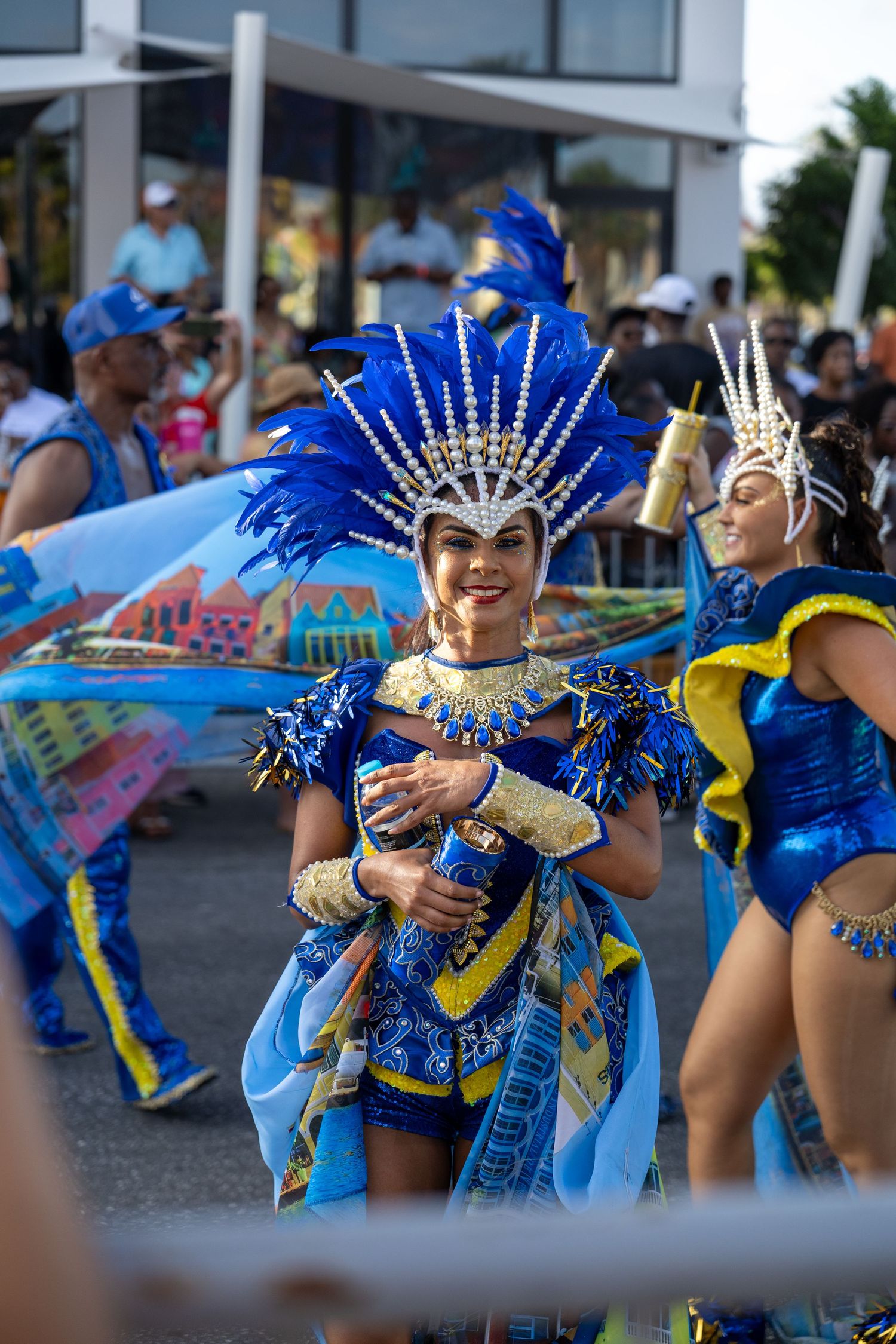 Dancers in sparkling blue and gold carnival attire perform synchronized movements at an outdoor event.