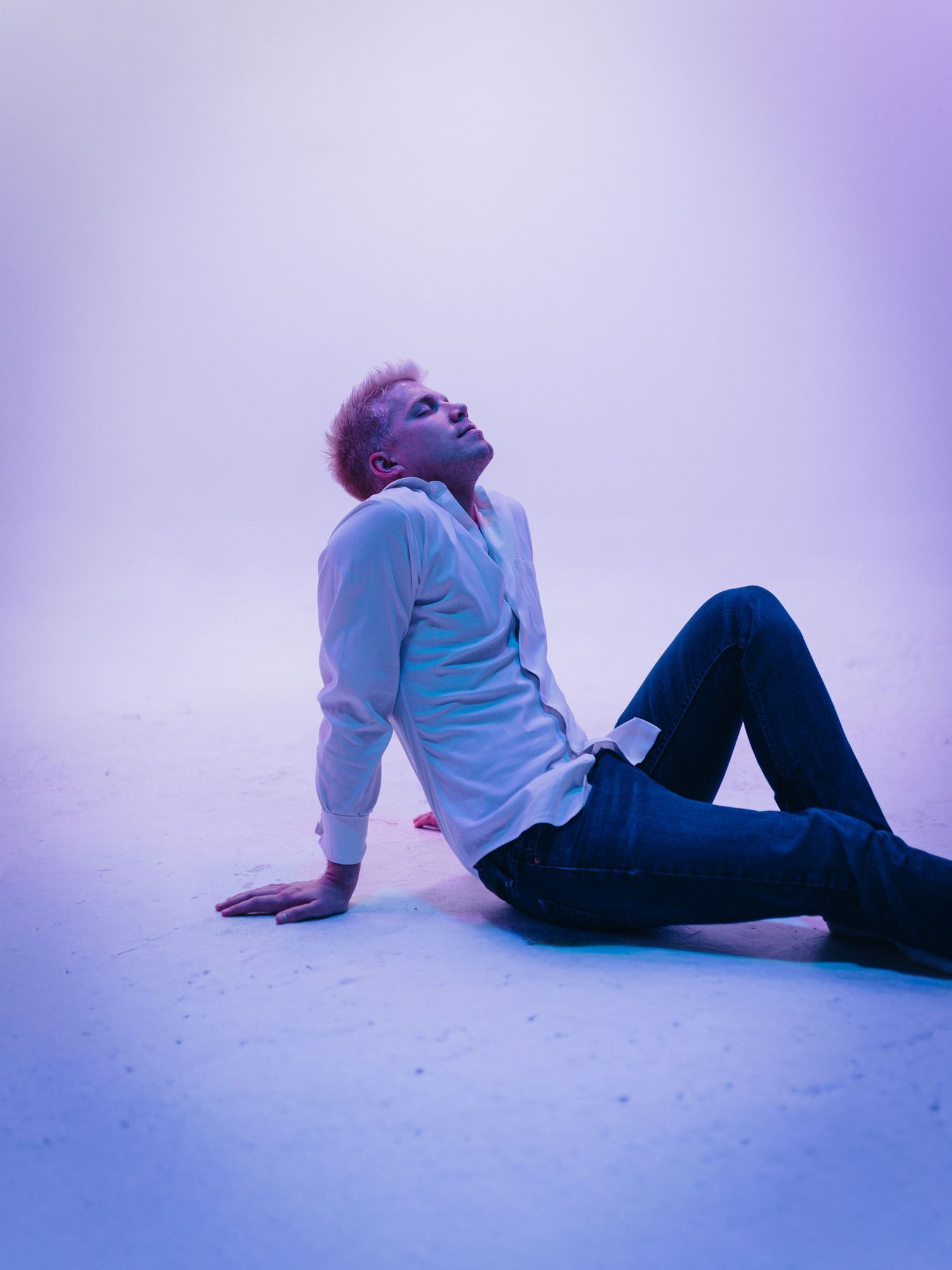 Young person with blonde hair sitting on floor in blue-purple light wearing white shirt and dark jeans.