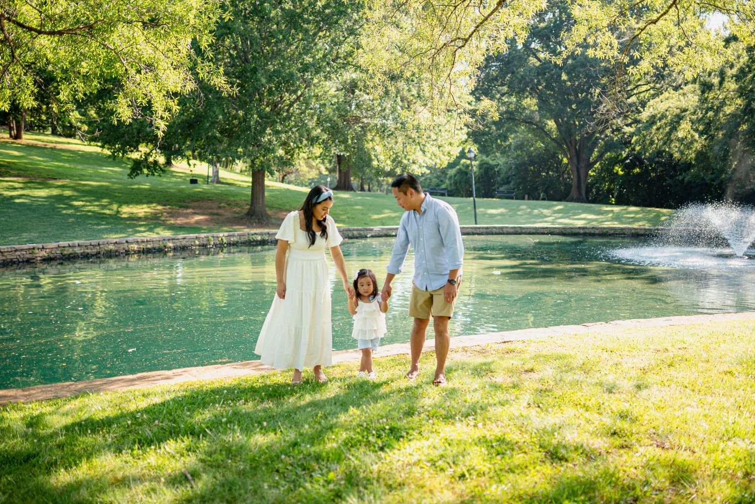 People walking along a grassy path near a lake with trees and fountain in the background.