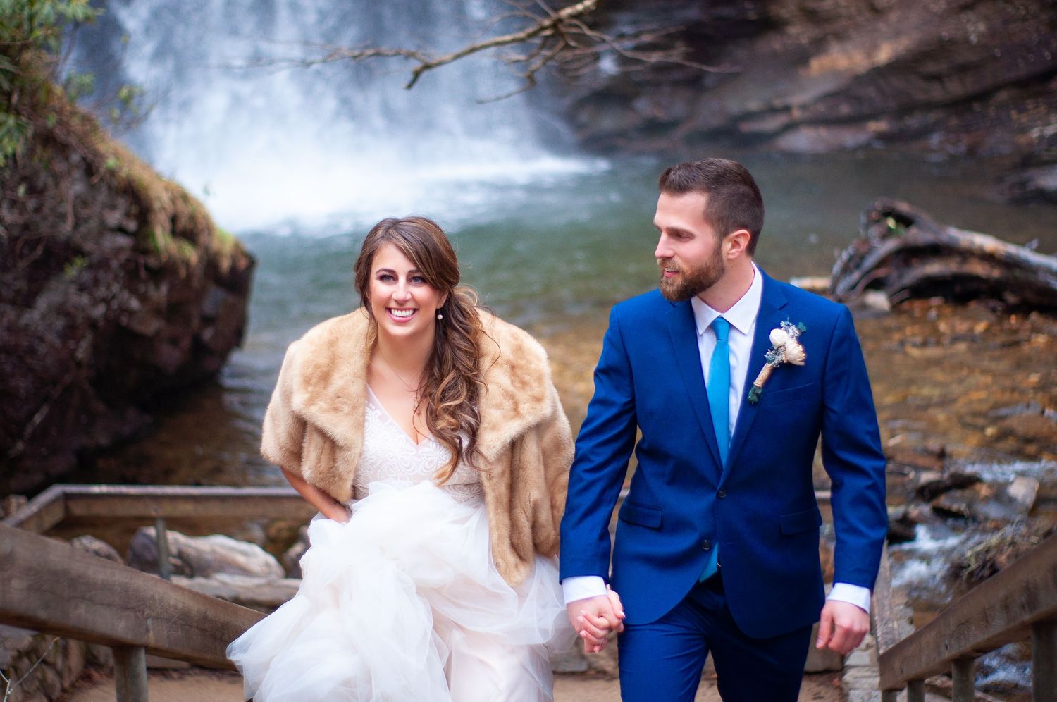 Newlyweds walking hand in hand near cascading waterfall.