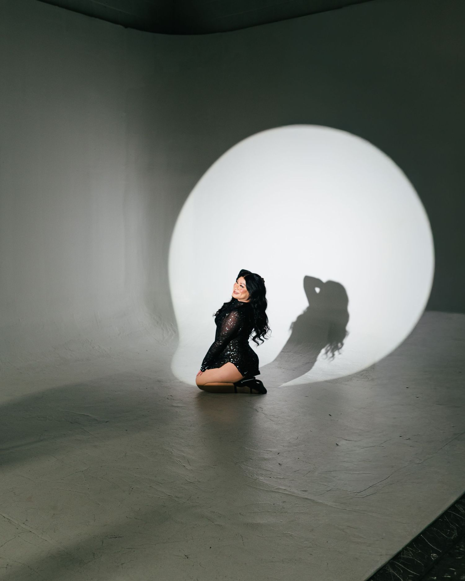 Woman in black lace bodysuit poses on floor before circular white light projection casting dramatic shadow on studio wall.