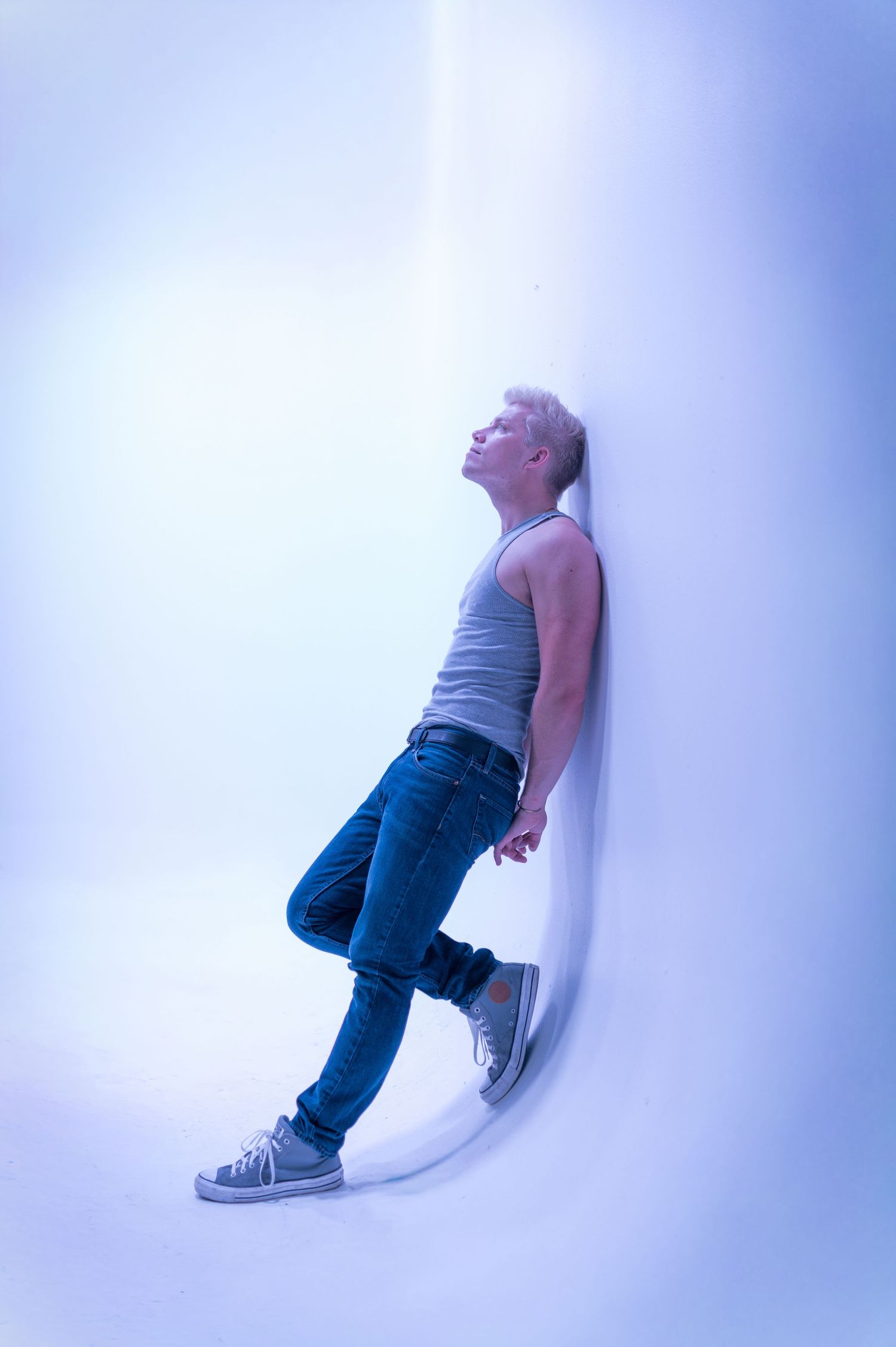 A person in jeans and tank top leans against a curved white wall under blue lighting, head tilted back.
