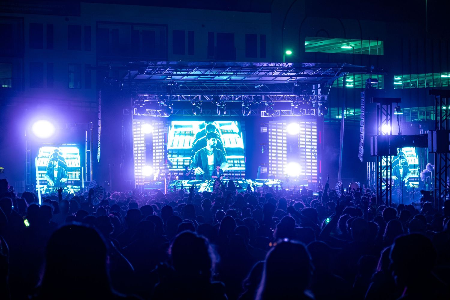 Outdoor concert at night with glowing blue and purple stage lights illuminating a large crowd of fans.
