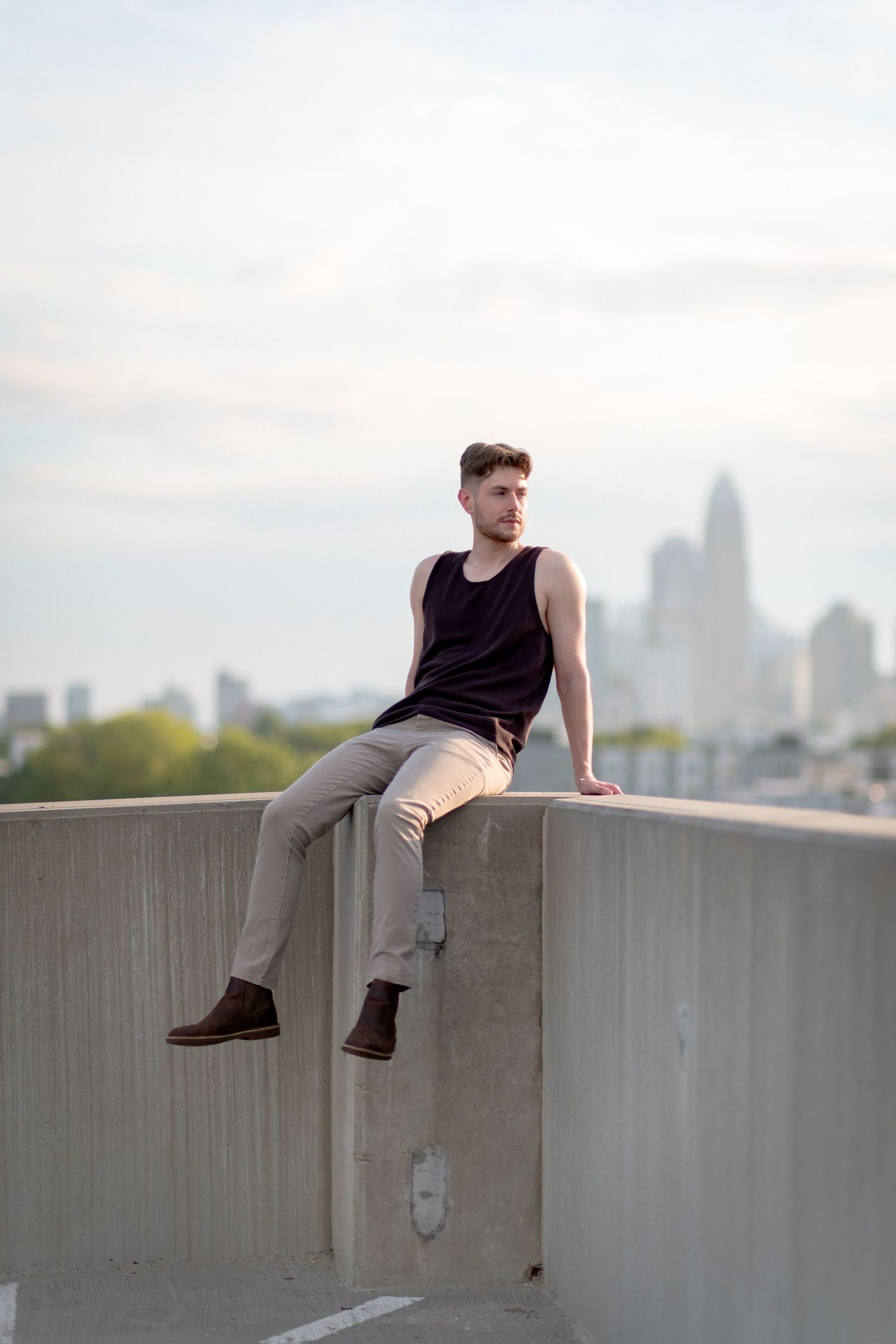 Young man in dark tank top and khakis sits on concrete ledge with city skyline backdrop.