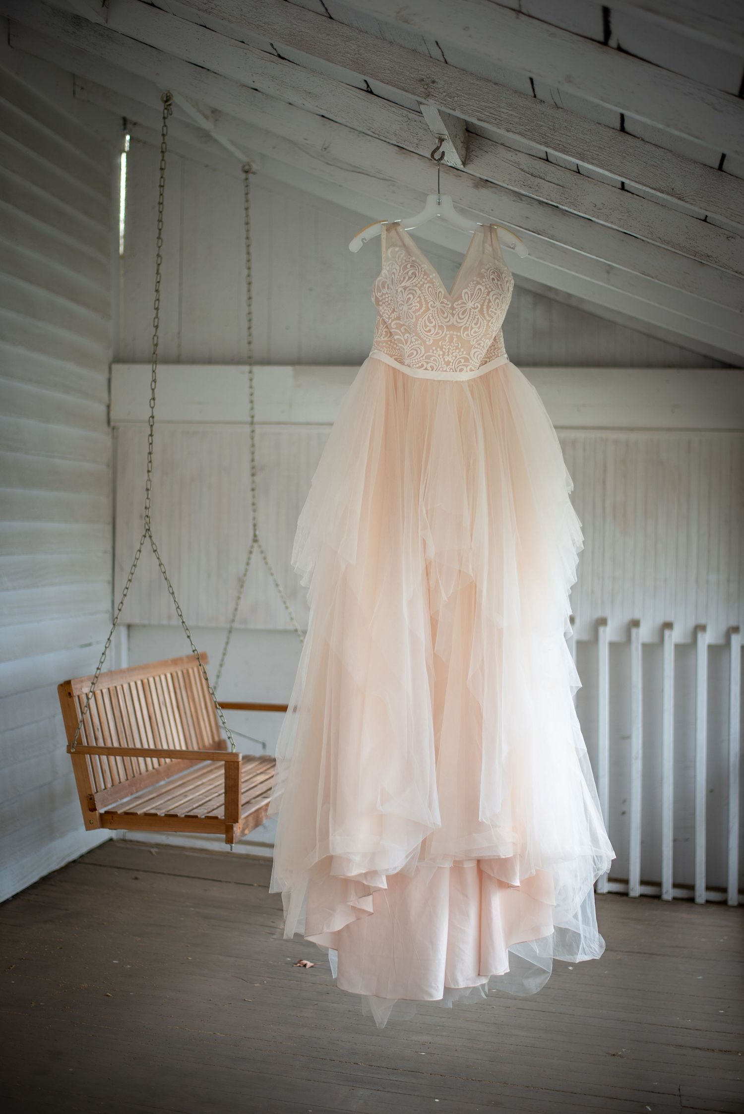 Blush pink wedding dress hanging on porch with wooden swing.